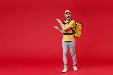Full length delivery employee man guy male 20s in yellow cap t-shirt uniform thermal food bag backpack work courier service during quarantine covid-19 virus, standing isolated on red background studio