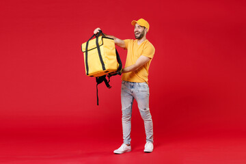 Full length delivery employee man guy male 20s in yellow cap t-shirt uniform thermal food bag backpack work courier service during quarantine covid-19 virus, standing isolated on red background studio - obrazy, fototapety, plakaty