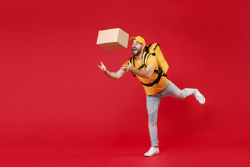Full length step delivery employee man male in yellow cap t-shirt uniform thermal food bag backpack hold cardboard box work courier service during quarantine covid-19 virus isolated on red background.