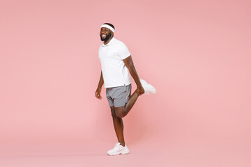 Full length portrait side view smiling young african american fitness sports man 20s in headband t-shirt doing stretching exercising for legs spending time in gym isolated on pink background studio.