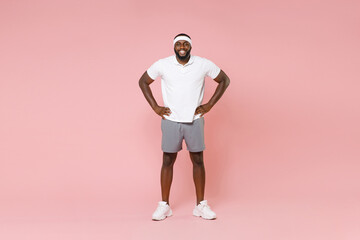 Fototapeta na wymiar Full length portrait of smiling young african american fitness sports man 20s in headband t-shirt standing with arms akimbo on waist spending time in gym isolated on pink color wall background studio.