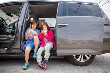 Two children sit side-by-side in an open van eating together