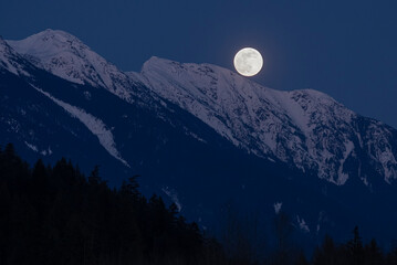 The full moon rises above snow covered mountain peaks on a spring night in the Coast Mountains of British Columbia.
