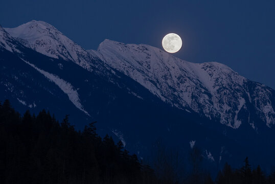The full moon rises above snow covered mountain peaks on a spring night in the Coast Mountains of British Columbia.