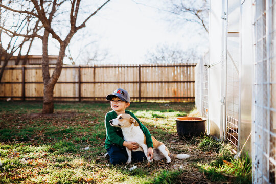 Young boy hugging corgi dog while sitting outside backyard greenhouse