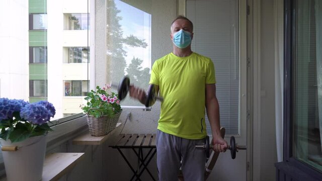 A man in sportswear playing sports with dumbbells on the balcony of his apartment during quarantine and self-isolation