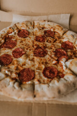 A large round juicy mouth-watering pizza with cheese and salami lies on a cardboard box from a delivery package. paper backing. close view. see flour. good thin dough