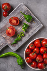 Fresh ripe tomatoes in bowl near to old wooden cutting board with hot chili peppers, stone concrete background, top view