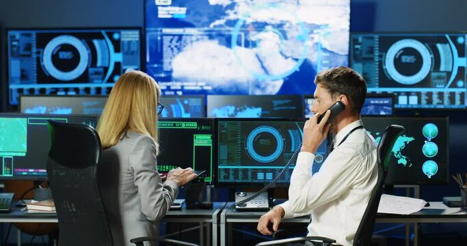 Rear on Caucasian male and female colleagues working in analytic digital military base. Woman using tablet device, tapping and scrolling. Man talking on telephone. Phone call. Back view on co-workers.