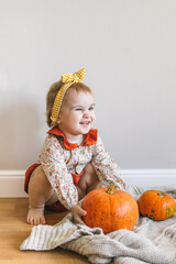 A one-year-old girl in a holiday outfit with a very surprised emotion on her face sits among the autumn decor and pumpkins