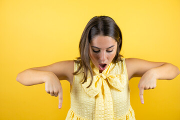 Young beautiful woman over isolated yellow background surprised, looking at the camera and pointing down with fingers and raised arms