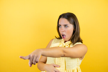 Young beautiful woman over isolated yellow background pointing with finger surprised ahead, open...