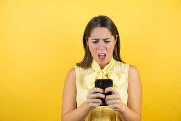 Young beautiful woman over isolated yellow background chatting with her phone and surprised