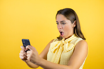 Young beautiful woman over isolated yellow background chatting with her phone and surprised