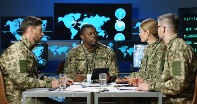 Mixed-races males and female militarian council sitting at table in uniforms, deciding war or defense tactics in dark monitoring room. Multiethnic men and woman officers talking, discussing terrorism.