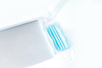 Modern laptop with a medical face mask and hand sanitizer bottle on top of a white desk.
