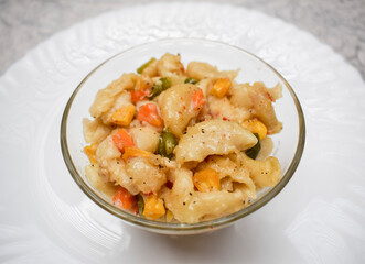 Close up of Vegetable macaroni cooked in White sauce served in a transparent bowl isolated on white background. Cooked pasta with carrot, corn, capsicum sprinkled with oregano and chilly flakes