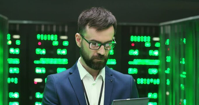 Caucasian handsome man in glasses standing in database room with servers, tapping on tablet device and browsing. Close up of male security worker in datacenter with working processors using gadget.