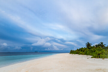 Tropical island shore with white sand and forest at sunset against sky with clouds