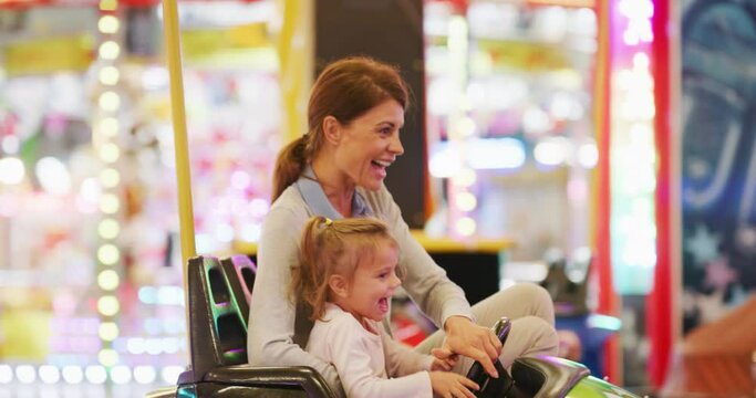 Authentic shot of a happy smiling family is having fun to drive and crush bumper cars at fun fair in amusement park with luna park lights at night.
