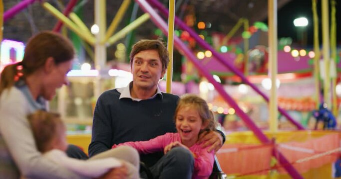 Authentic shot of a happy smiling father and his little daughter are having fun to eat together a cotton candy in amusement park with luna park lights at night.