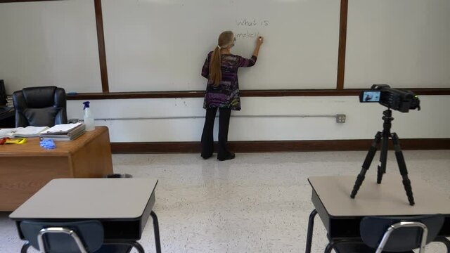 Camera pushes in on female civics or grammar teacher wearing medical face mask, writing what is implicit bias on a white board in a school classroom. Concept posing question for students.