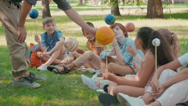 Medium shot of unrecognizable male science teacher talking to cute schoolchildren sitting on blankets in park and holding models of planets while learning about solar system