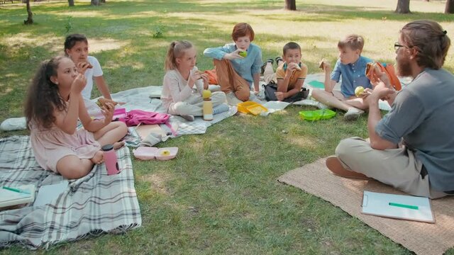 Slowmo tracking of bearded male teacher and his little students sitting on blankets on green grass in park on summer day and eating lunch