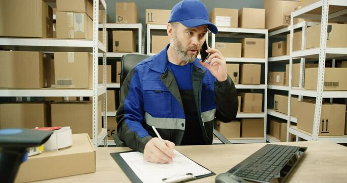 Caucasian man talking on mobile phone while working at post office store with parcels and on computer. Postman registering box and filling in invoice while speaking on cellphone in mail storage.