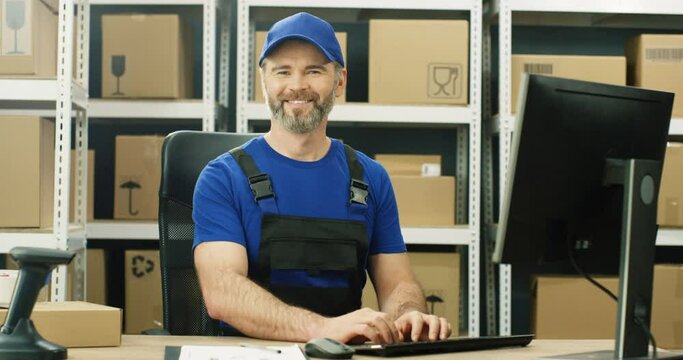 Portrait shot of cheerful Caucasian postman in iniform and cap sitting at table in postal office store and working at computer. Handsome male courier smiling happily to camera in storage of parcels.