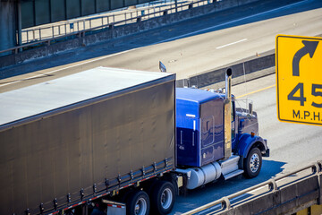 Bright blue big rig semi truck transporting commercial cargo in covered semi trailer with frame driving on the interstate highway