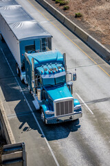 Classic big rig blue semi truck tractor transporting cargo in bulk semi trailer with covered top driving on the  elevated road