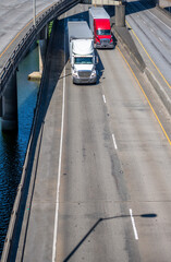 Different two big rigs semi trucks with semi trailers running side by side on the overpass road intersection along the river