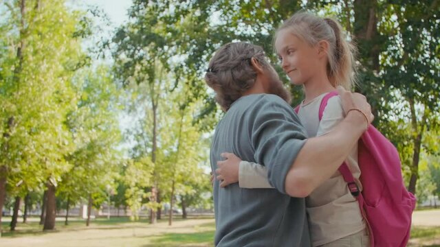 Medium shot of bearded father smiling and hugging cute schoolgirl with backpack, then waving her goodbye
