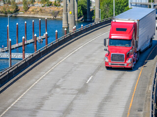 Red big rig semi truck with reefer semi trailer running on the turning overpass road intersection along the river with pier