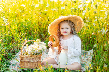 beautiful little girl sitting in a straw hat in a yellow field with wild flowers with a bottle of milk and a bagel, picnic in the field