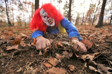 Halloween holiday. A scary evil clown crawls through the autumn foliage .Halloween carnival party outdoors.Masquerades and festive carnivals in October.Horror and fear.