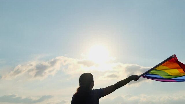 Rainbow LGBT flag on sun sky. Silhouette woman holding gay pride lgbt flag in hands - lesbian, gay, bisexual, transgender social movements, Concept of happiness freedom love same-sex couple 4 K slowmo