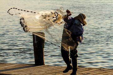 A fisherman wearing overalls and boots as well as a bucket hat is throwing a cast net into the sea...