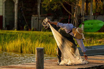 A fisherman wearing overalls, plastic boots and a bucket hat is preparing his cast net before throwing it into the Chesapeake Bay at sunset. He is on a wooden pier and sun reflects from water surface.