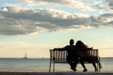 A young African American couple is sitting on a bench looking at sea and Chesapeake Bay bridge. Man...