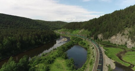 Freight long train carries with coal carriages an electric locomotive by two-sided Trans Siberian railways under the rock and near mountains river / Aerial drone wide view at summer sunny day