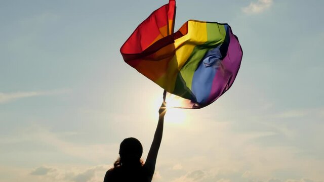 Rainbow LGBT flag on sun blue sky. Female woman holding gay pride flag in hands -lesbian, gay, bisexual, transgender social movements. Concept of happiness freedom love same-sex couple, slow-mo