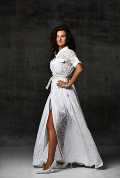 Elegant powerful brunette woman in white long lace dress with short sleeves and high heeled shoes stands looking at camera