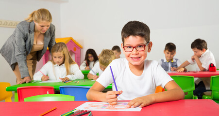 Little schoolboy drawing with color pencils at desk in classroom