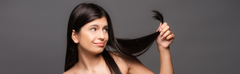 Naklejka premium naked skeptical brunette long haired woman looking at split ends isolated on black, panoramic shot