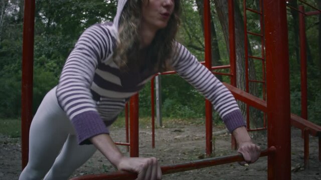 Young caucasian woman doing sports on the playground in the forest on a cloudy day. A woman pulls herself up on a horizontal bar.