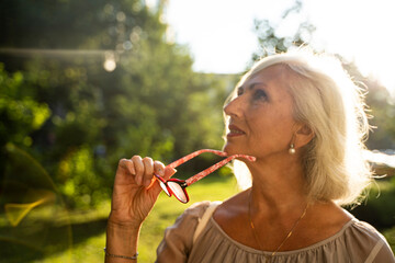 blurred portrait of pensive gray-haired woman takes off her glasses and looks up while on the street in summer.