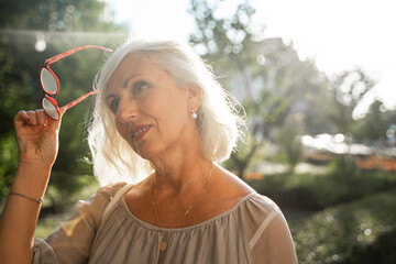 pensive gray-haired woman takes off her glasses and looks away while on the street in summer.