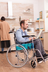 Handicapped entrepreneur in wheelchair working on laptop while wife is preparing meal in kitchen....